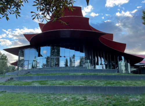 Modernes Rundgebäude mit geschwungenen roten Dachteilen, Glasfassade, Stufen und Rasen, blauer Himmel